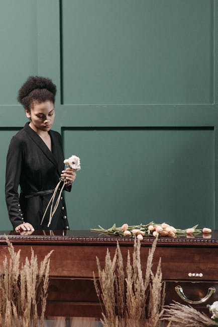 A young woman with natural curly hair dressed in a black coat stands beside a wooden casket, holding a single white flower. In front of her, on the casket's surface, lies an arrangement of pale pink r