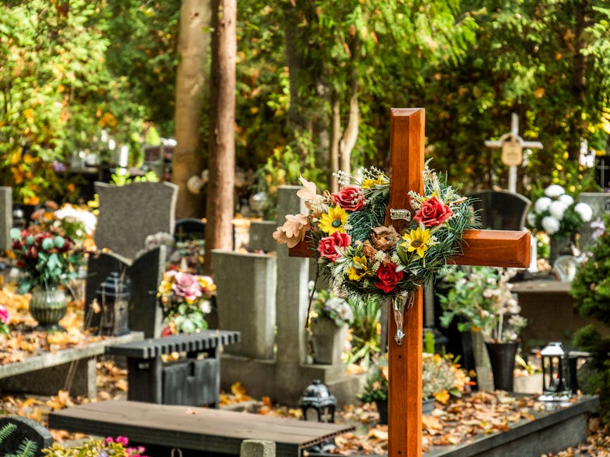 A wooden funeral cross adorned with a floral arrangement featuring red roses, yellow daisies, beige lisianthus, and sprigs of greenery, positioned among gravestones and memorial plaques in a cemetery 