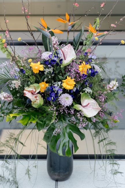 A vibrant floral arrangement in a tall, dark ceramic vase, featuring a mix of fresh flowers including white anthuriums with pink accents, bright yellow daisies, deep blue hyacinths, and delicate purpl