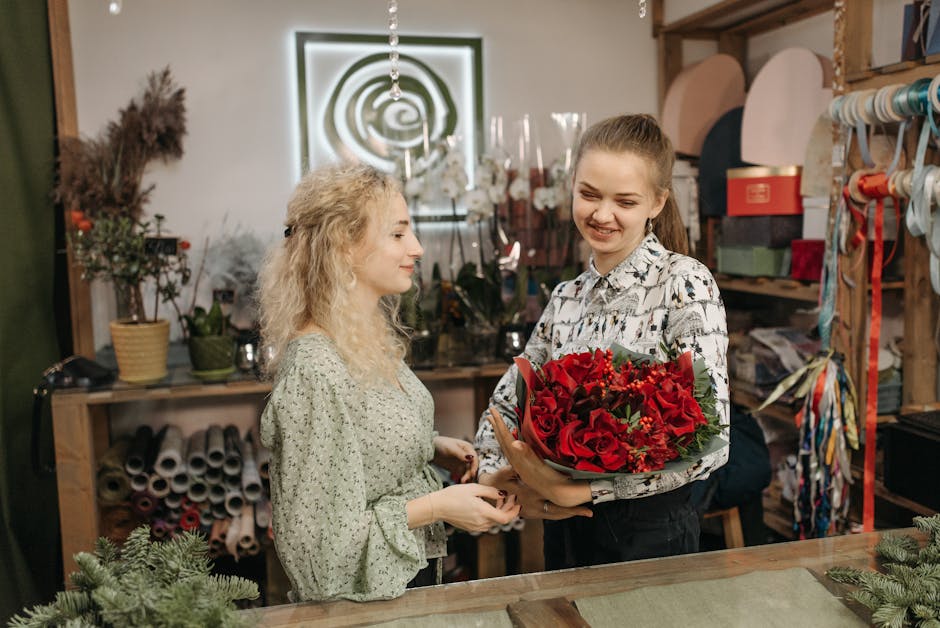 A young woman with light brown hair tied back, wearing a patterned long-sleeve blouse, is in the process of handing over a large bouquet of fresh red roses, red hypericum berries, and green foliage, w