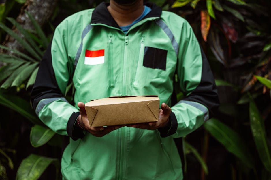 A person wearing a green and black jacket, with an Indonesian flag patch on the chest, holding a plain brown cardboard box with their hands. The background is filled with lush, green tropical foliage,