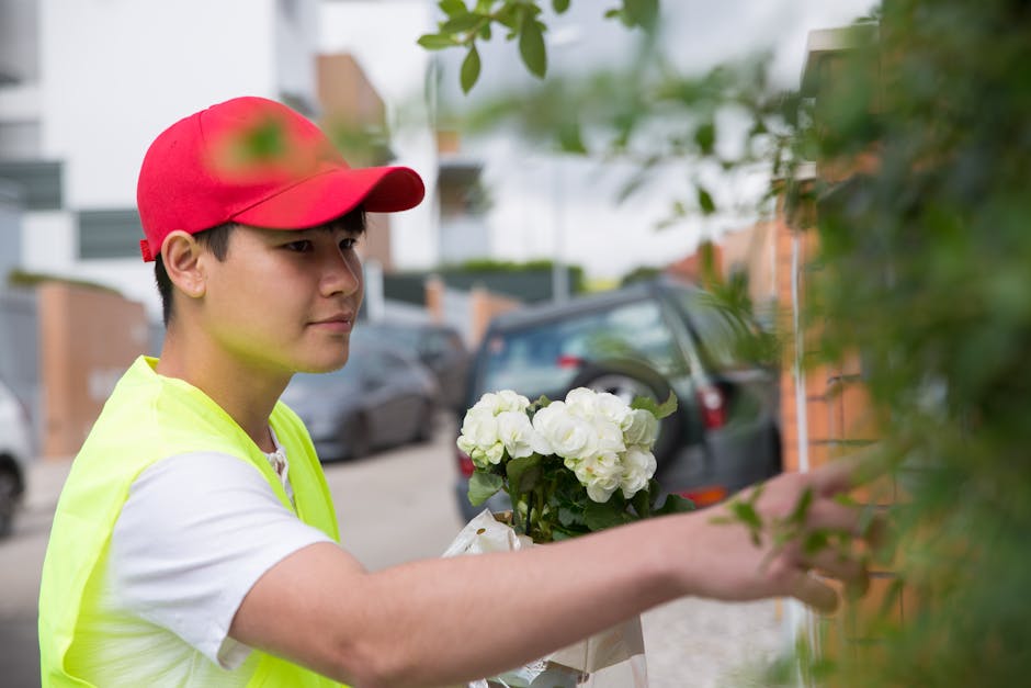 A young male florist wearing a red cap and a high-visibility yellow vest arranges a floral arrangement outdoors. He is holding a bouquet of white hydrangeas and is carefully placing it against a green