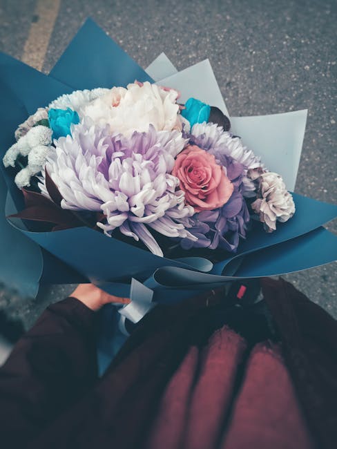 A hand holding a beautifully arranged floral bouquet featuring large white and purple chrysanthemums, a blush pink rose, and smaller purple and white blooms, all wrapped in elegant blue and white pape