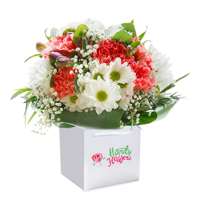 A strikingly lush bouquet, shaped to perfection and standing upright in a modern white gift bag, brings to mind celebrations along Station Road or quiet moments at Chingford's Queen Elizabeth's Hunting Lodge. The star of the composition is a blend of soft coral and vivid red carnations, their luxuriously ruffled petals clustered thickly to form an abundant core. Bright, crisp white daisies with cheerful chartreuse centres provide a lively counterpoint, their petals catching the light as if basking in a window overlooking Chingford Green. Between these main blooms, delicate sprays of white baby's breath weave, infusing the display with a subtle, airy grace and timeless elegance. Along one side, a hint of unopened buds, tinged with lavender and wine, peeks through-suggesting the unfolding promise of spring in local gardens. All is held together by an embrace of robust, glossy green foliage that arches protectively around the arrangement's base, offering visual structure and balance. The gift bag provides easy, stylish portability, perfect for hand delivery across Chingford, while its minimalist design ensures all attention remains on the floral beauty within. This bouquet feels both generously abundant and meticulously composed, an ideal gesture for milestone birthdays, anniversary dinners, or simple moments of appreciation in the heart of the community, crafted with the local sensibility and care Chingford Flowers is known for.