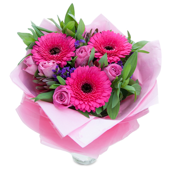 This arrangement combines playfulness and grace: three saturated fuchsia Gerbera daisies set a bold, cheerful tone with their radiating petals and dark central discs, while nearby lavender-pink roses soften the palette with tightly furled, velvety layers that promise a gentle, classic fragrance. Small clusters of purple-blue statice float between the larger blooms, introducing a paper-like texture and a hint of the countryside tucked into an urban bouquet - a detail that brings to mind wildflowers edging the paths of Epping Forest. Broad, glossy foliage, suggestive of salal or eucalyptus, frames and anchors the floral shapes, creating depth and a fresh green contrast that makes the pinks sing. The flowers are lovingly wrapped: an inner cushion of soft pale pink tissue creates warmth and softness at the base, while an outer veil of translucent fuchsia-pink cellophane adds a subtle sheen and ties the colour story together; the careful folds reveal the top rim of a clear glass vase, indicating the bouquet is prepared for immediate display. In soft daylight the colours glow; the composition reads as an intimate, celebratory gift, perfectly suited for a birthday, an anniversary, or a tender 'just because' surprise after a stroll past Queen Elizabeth's Hunting Lodge or a morning at Chingford Mount - a bouquet designed to bring comfort, joy and the scent of fresh flowers into any home.