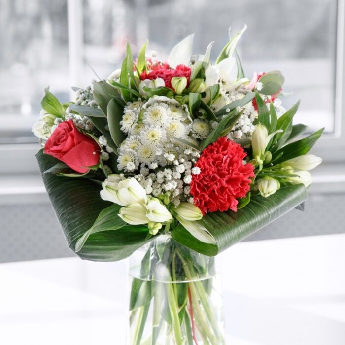 A contemporary yet timeless bouquet stands in a simple clear glass cylinder vase, its clean lines allowing the flowers to command full attention. Below the waterline, smooth, straight stems form a tidy green framework, evidence of a florist's careful preparation. Above, the composition unfolds in a harmonious dance of red, white, and green. Deep red roses, their petals velvety and full of life, gather near the centre, while a single large crimson carnation, densely ruffled, anchors the front with sculptural detail. Around these, white and cream blooms offer a softer counterpoint: white alstroemeria with slender petals and subtle speckles, rounded daisy-like flowers with sunny centres, and sleek white lily buds yet to open, hinting at unfolding beauty over the coming days. Fine sprays of gypsophila weave through the bouquet, each cluster of tiny white flowers adding a sense of airiness and gentle celebration. The whole arrangement is framed by lush foliage in multiple shades of green. Broad, glossy leaves are wrapped and curled to form a polished collar at the base, while longer, pointed greenery and eucalyptus-style stems extend upwards, creating depth, rhythm, and a gentle sense of movement. The bouquet sits on a pristine white surface, in front of a softly blurred, bright background that could easily be a Chingford home bathed in daylight or a light-filled corner of a local café near Station Road. Soft, even light caresses the petals and leaves, emphasising freshness and craft. It feels like a thoughtfully arranged centrepiece, perfect for bringing warmth and gratitude to a family gathering, celebrating friendship, or marking a quiet yet meaningful moment in everyday Chingford life.