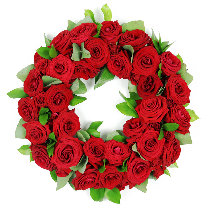 An eloquent, deeply symbolic wreath of dozens of vibrant red roses sits against a pristine white background, its circular form unbroken and serene. The blooms vary from tightly closed buds to fully opened petals, each rose in a shade of saturated crimson that suggests both passion and reverence. Interspersed throughout the wreath are clusters of fresh green foliage-broad, matte leaves reminiscent of eucalyptus and sharper, glossy ruscus-that create a lively yet refined texture and a striking colour counterpoint. The arrangement is intentionally full, with petals and leaves layering over one another to give a three-dimensional sense of depth. This wreath speaks of memorials, respect, and heartfelt condolence, and is a powerful visual element for articles about funeral arrangements, anniversaries, or commemorations. The stark white background enhances the solemn beauty and helps the red glow feel all the more vivid. Prepared by a local Chingford florist, it carries the hallmarks of careful craftsmanship: balanced spacing, even bloom distribution, and a natural, graceful curve. It can be displayed at church entrances, funeral homes, or at family tributes, where its timeless elegance supports quiet reflection. Available for delivery across Chingford and its environs, this piece offers immediate reassurance and a dignified way to express sympathy to loved ones near and far.
